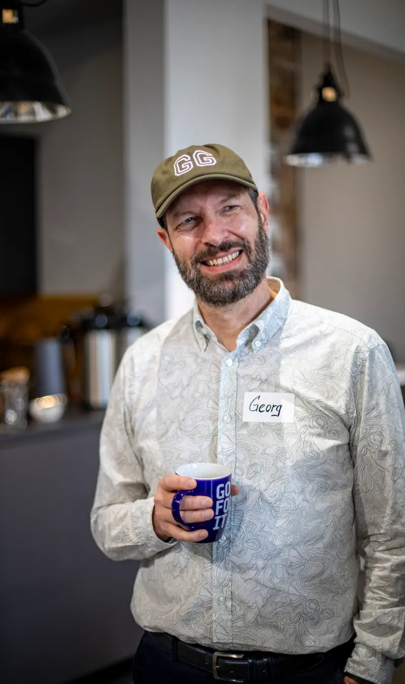 Georg Fischer hält mit Kappe und Namensschild eine dunkelblaue Tasse mit der Aufschrift GO FOR IT.